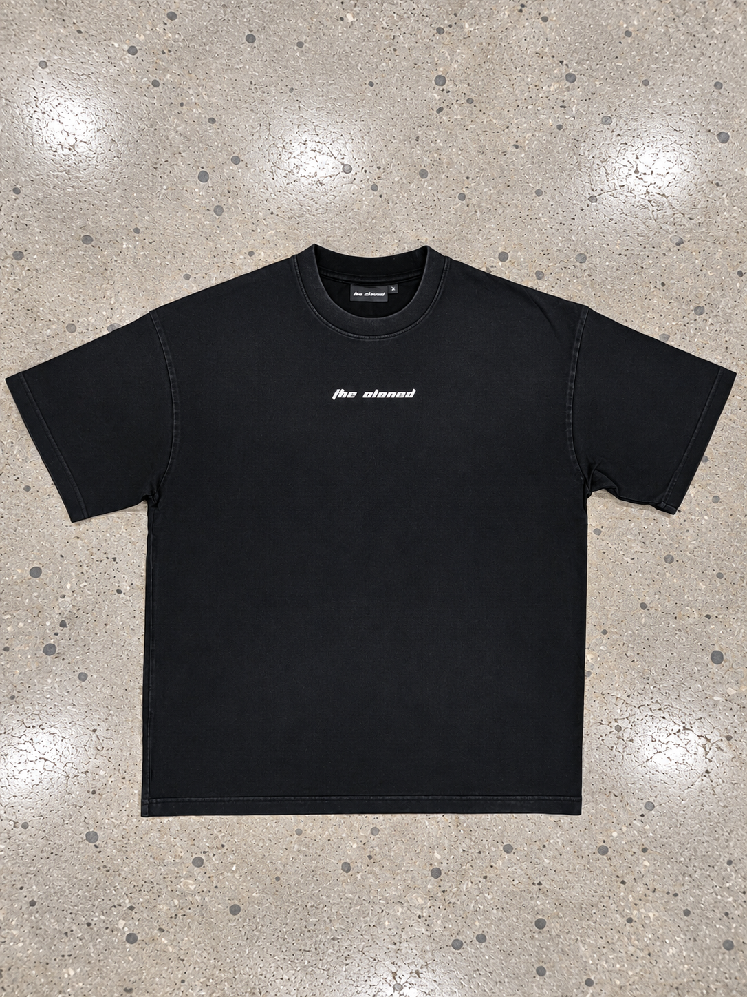 A black short-sleeve t-shirt with a small white vertical logo on the left chest, laid flat on a speckled concrete floor.